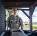 A U.S. Army Military Police soldier, identified by the name tape 'Berger,' stands at what appears to be a checkpoint or guard post wearing full tactical gear including a plate carrier vest with 'MILITARY POLICE' markings. The soldier is in OCP (Operational Camouflage Pattern) uniform and appears to be on duty at an outdoor installation, with a military vehicle (HMMWV/Humvee) visible in the background. The setting suggests a forward operating base or training area in a temperate region.