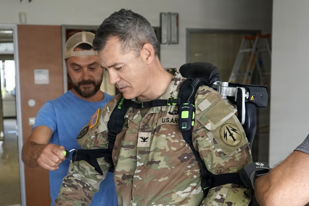 A U.S. Army Colonel in OCP (Operational Camouflage Pattern) uniform is being fitted with what appears to be a powered exoskeleton or advanced load-bearing equipment by a civilian technician in a blue shirt. The military officer's rank insignia (eagle/full bird colonel) is visible on his chest, along with a U.S. Army tape and unit patches. The setting appears to be an indoor facility, possibly a demonstration or testing environment.