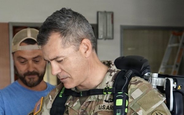 A U.S. Army Colonel in OCP (Operational Camouflage Pattern) uniform is being fitted with what appears to be a powered exoskeleton or advanced load-bearing equipment by a civilian technician in a blue shirt. The military officer's rank insignia (eagle/full bird colonel) is visible on his chest, along with a U.S. Army tape and unit patches. The setting appears to be an indoor facility, possibly a demonstration or testing environment.