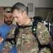 A U.S. Army Colonel in OCP (Operational Camouflage Pattern) uniform is being fitted with what appears to be a powered exoskeleton or advanced load-bearing equipment by a civilian technician in a blue shirt. The military officer's rank insignia (eagle/full bird colonel) is visible on his chest, along with a U.S. Army tape and unit patches. The setting appears to be an indoor facility, possibly a demonstration or testing environment.