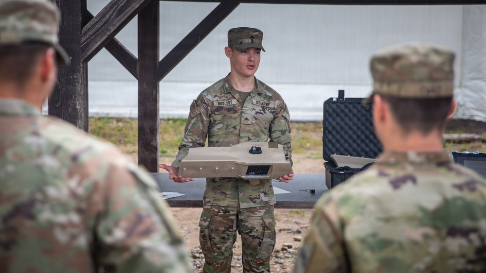 A U.S. Army soldier identified by name tape as 'GOOLSBY' is conducting a briefing or equipment demonstration while holding a flat rectangular device, likely a piece of military equipment or controller. He is addressing at least two other soldiers whose backs are visible in the foreground. The setting appears to be an outdoor covered area or pavilion with a table and a hard-shell equipment case visible in the background.