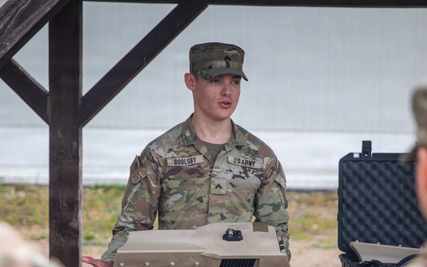 A U.S. Army soldier identified by name tape as 'GOOLSBY' is conducting a briefing or equipment demonstration while holding a flat rectangular device, likely a piece of military equipment or controller. He is addressing at least two other soldiers whose backs are visible in the foreground. The setting appears to be an outdoor covered area or pavilion with a table and a hard-shell equipment case visible in the background.