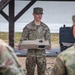 A U.S. Army soldier identified by name tape as 'GOOLSBY' is conducting a briefing or equipment demonstration while holding a flat rectangular device, likely a piece of military equipment or controller. He is addressing at least two other soldiers whose backs are visible in the foreground. The setting appears to be an outdoor covered area or pavilion with a table and a hard-shell equipment case visible in the background.