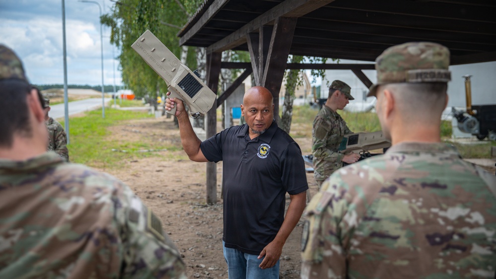 A civilian instructor in a black polo shirt holds up a military equipment component while briefing a group of soldiers in OCP (Operational Camouflage Pattern) uniforms outdoors. The setting appears to be a training area or military installation, with a covered shelter structure visible in the background. Additional soldiers can be seen working with equipment in the background.
