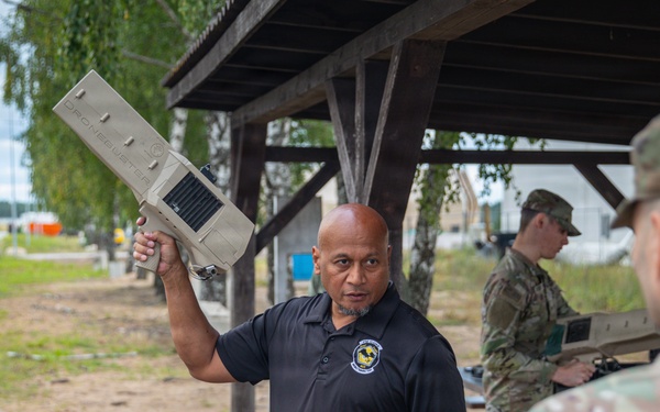 A civilian instructor in a black polo shirt holds up a military equipment component while briefing a group of soldiers in OCP (Operational Camouflage Pattern) uniforms outdoors. The setting appears to be a training area or military installation, with a covered shelter structure visible in the background. Additional soldiers can be seen working with equipment in the background.