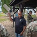 A civilian instructor in a black polo shirt holds up a military equipment component while briefing a group of soldiers in OCP (Operational Camouflage Pattern) uniforms outdoors. The setting appears to be a training area or military installation, with a covered shelter structure visible in the background. Additional soldiers can be seen working with equipment in the background.
