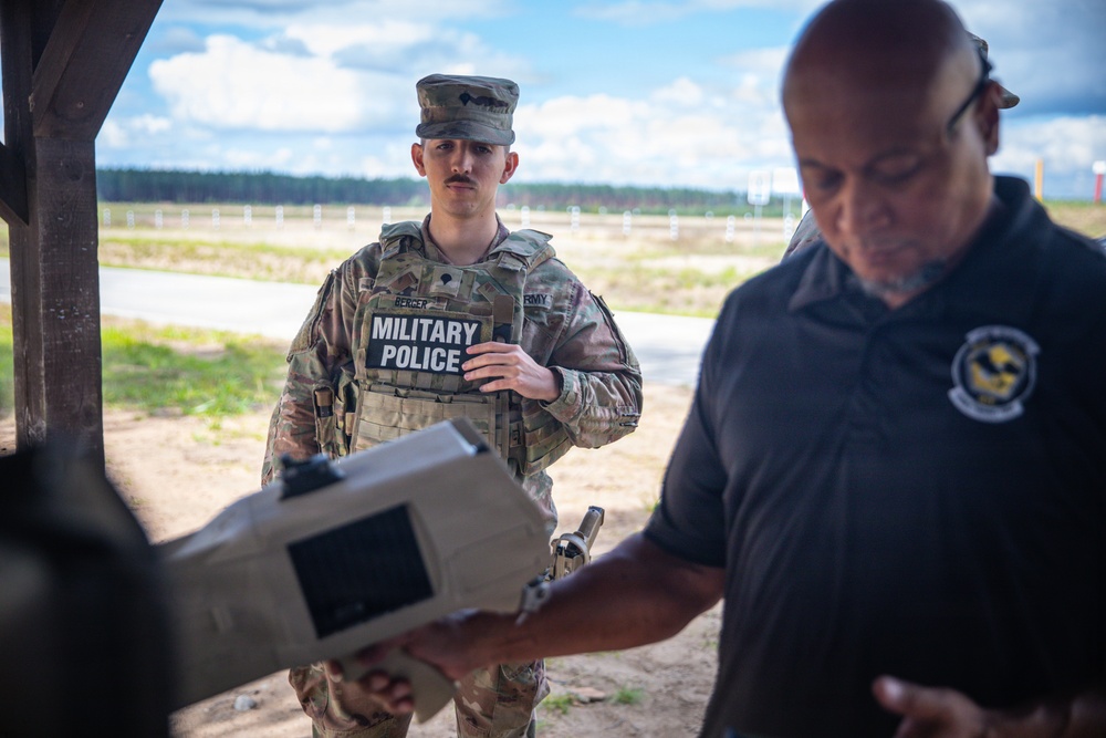 A U.S. Army Military Police soldier in full combat uniform and body armor stands at what appears to be an outdoor shooting range or training facility. A civilian or contractor in a dark polo shirt is handling a piece of equipment in the foreground. The setting suggests a training or demonstration exercise.