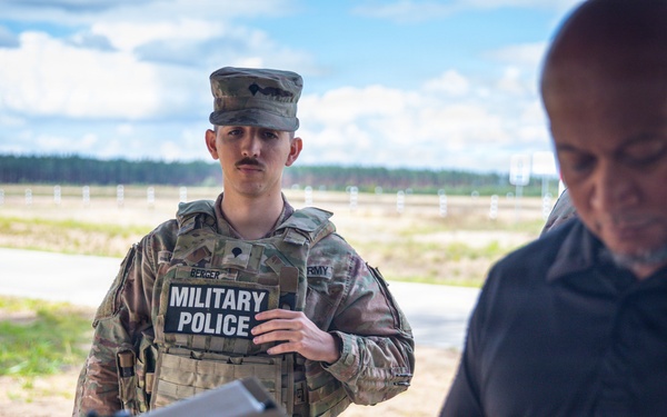 A U.S. Army Military Police soldier in full combat uniform and body armor stands at what appears to be an outdoor shooting range or training facility. A civilian or contractor in a dark polo shirt is handling a piece of equipment in the foreground. The setting suggests a training or demonstration exercise.