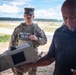A U.S. Army Military Police soldier in full combat uniform and body armor stands at what appears to be an outdoor shooting range or training facility. A civilian or contractor in a dark polo shirt is handling a piece of equipment in the foreground. The setting suggests a training or demonstration exercise.