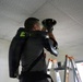 A male technician or installer is standing on a ladder working on a ceiling fixture, likely installing or servicing a light or camera in a drop ceiling. He is wearing a black exoskeleton support device on his back and torso, designed to reduce strain during overhead work. The setting appears to be a commercial or institutional interior space.