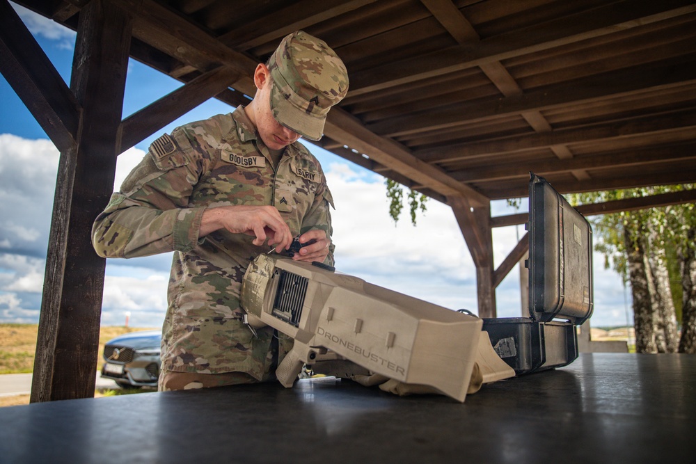 A U.S. Army soldier identified by name tape as 'GOOLSBY' is working on a DroneBuster counter-drone device on a table under a wooden shelter. The soldier is wearing OCP (Operational Camouflage Pattern) uniform and appears to be assembling or configuring the anti-drone equipment. A hard-shell transport case is visible beside the device, suggesting recent unpacking or preparation for use.