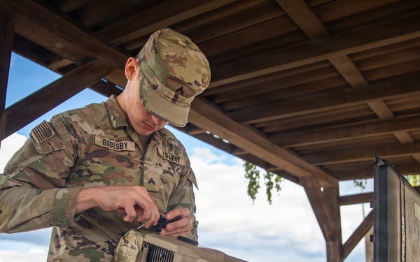 A U.S. Army soldier identified by name tape as 'GOOLSBY' is working on a DroneBuster counter-drone device on a table under a wooden shelter. The soldier is wearing OCP (Operational Camouflage Pattern) uniform and appears to be assembling or configuring the anti-drone equipment. A hard-shell transport case is visible beside the device, suggesting recent unpacking or preparation for use.