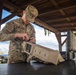 A U.S. Army soldier identified by name tape as 'GOOLSBY' is working on a DroneBuster counter-drone device on a table under a wooden shelter. The soldier is wearing OCP (Operational Camouflage Pattern) uniform and appears to be assembling or configuring the anti-drone equipment. A hard-shell transport case is visible beside the device, suggesting recent unpacking or preparation for use.