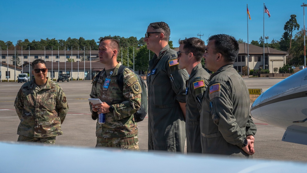 A group of five U.S. military personnel are standing on an aircraft ramp or flight line, appearing to observe or receive a briefing near an aircraft. Two individuals are wearing Army Combat Uniforms (ACU/OCP pattern) while three are wearing olive drab flight suits with U.S. flag patches. The setting appears to be a military air base, with flagpoles visible in the background flying what appear to be U.S. and German flags.