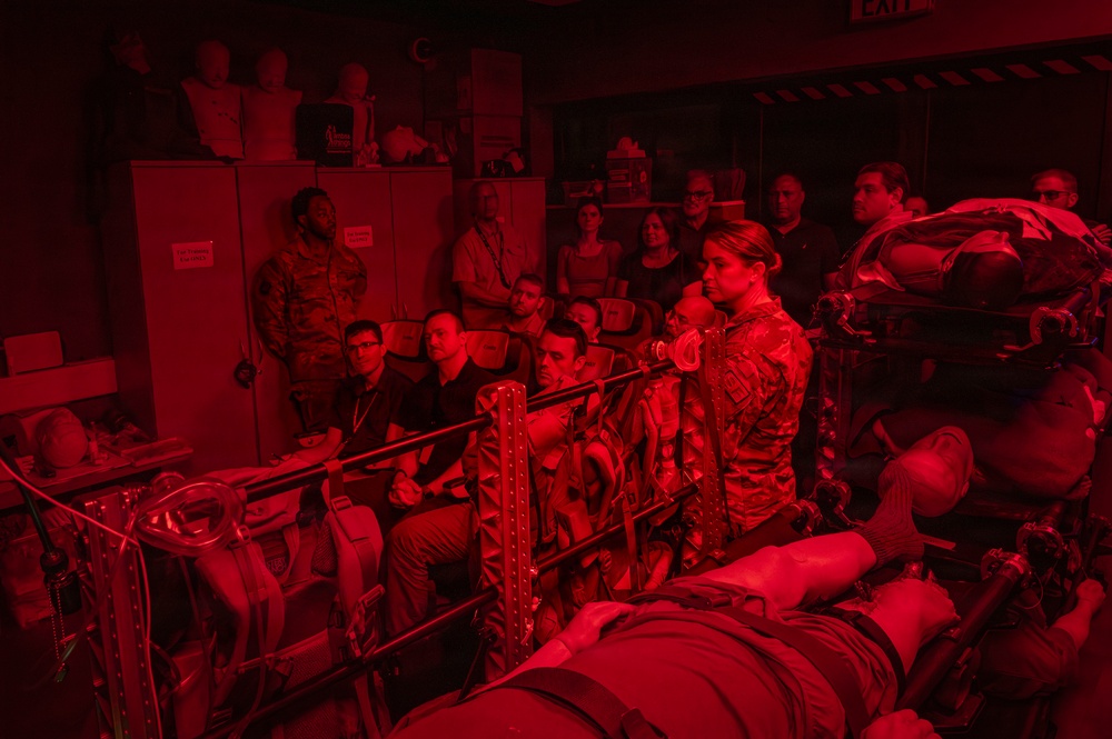 A group of people are gathered in a room bathed in red lighting, observing what appears to be a military or aerospace medical training demonstration. A person in camouflage uniform stands near equipment that resembles litter/stretcher systems used for aeromedical evacuation. Several observers, including uniformed military personnel and civilians with lanyards/badges, are watching attentively.