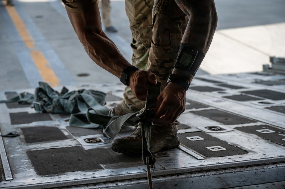 A military personnel member in camouflage uniform is working on the cargo floor of what appears to be a military transport aircraft, handling tie-down straps or cargo restraint equipment. The floor features the characteristic anti-skid pads and tie-down rings typical of military cargo aircraft such as a C-17 or C-130. The close-up perspective emphasizes the hands-on nature of cargo loading or securing operations.