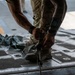 A military personnel member in camouflage uniform is working on the cargo floor of what appears to be a military transport aircraft, handling tie-down straps or cargo restraint equipment. The floor features the characteristic anti-skid pads and tie-down rings typical of military cargo aircraft such as a C-17 or C-130. The close-up perspective emphasizes the hands-on nature of cargo loading or securing operations.