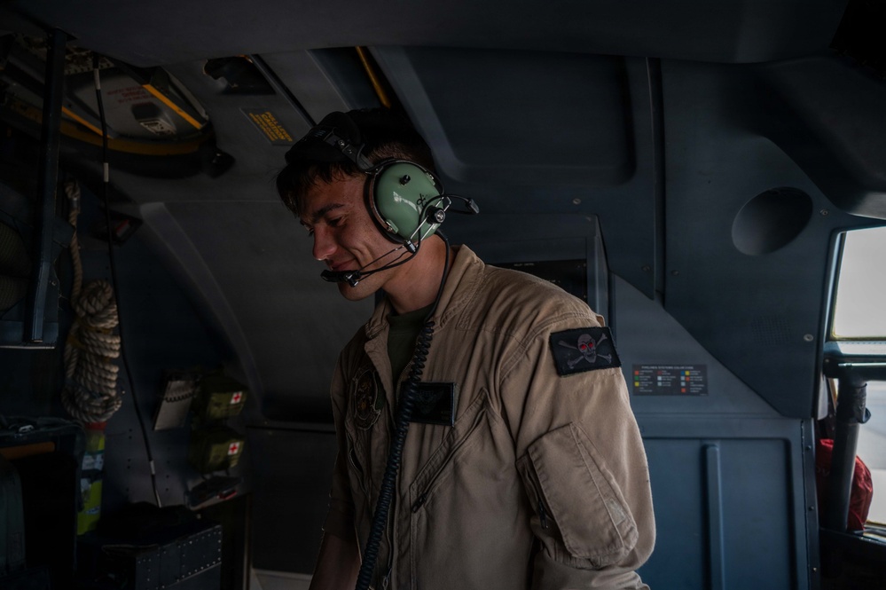 A military aircrew member is photographed inside what appears to be a military transport aircraft or helicopter. He is wearing a tan/khaki flight suit with a skull-and-crossbones patch and a green aviation communications headset with boom microphone. The image is dramatically lit with strong shadows, giving it a documentary or editorial quality.