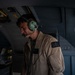A military aircrew member is photographed inside what appears to be a military transport aircraft or helicopter. He is wearing a tan/khaki flight suit with a skull-and-crossbones patch and a green aviation communications headset with boom microphone. The image is dramatically lit with strong shadows, giving it a documentary or editorial quality.