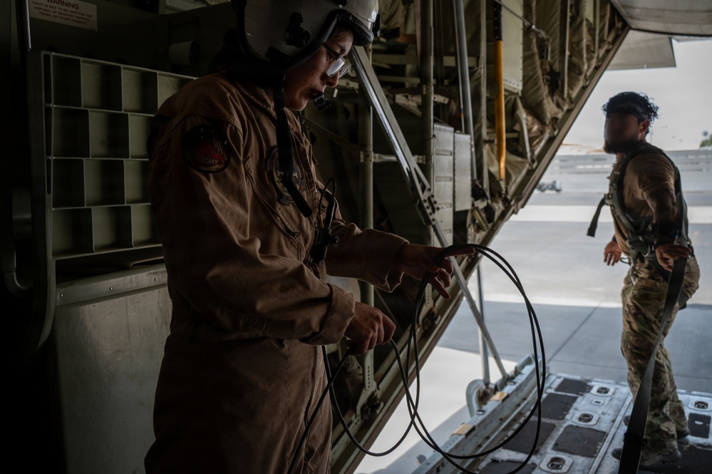 Two military personnel are working inside and near the rear cargo ramp of a large military transport aircraft, likely a C-130 Hercules. The foreground figure, wearing a flight suit and helmet, is handling a coiled cable or cord. A second individual in camouflage uniform stands near the open ramp, with an airfield visible in the background.