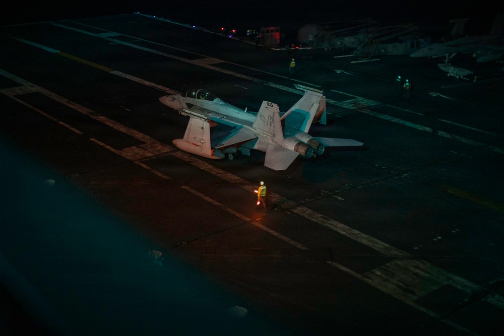 A nighttime scene on an aircraft carrier flight deck showing a military jet aircraft, likely an F/A-18 Hornet or Super Hornet, parked on the deck. Personnel in reflective safety gear are visible on the deck, with one individual prominently illuminated by orange light in the foreground. The image is taken from an elevated angle, revealing the dark expanse of the carrier deck and additional personnel and equipment in the background.