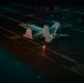 A nighttime scene on an aircraft carrier flight deck showing a military jet aircraft, likely an F/A-18 Hornet or Super Hornet, parked on the deck. Personnel in reflective safety gear are visible on the deck, with one individual prominently illuminated by orange light in the foreground. The image is taken from an elevated angle, revealing the dark expanse of the carrier deck and additional personnel and equipment in the background.