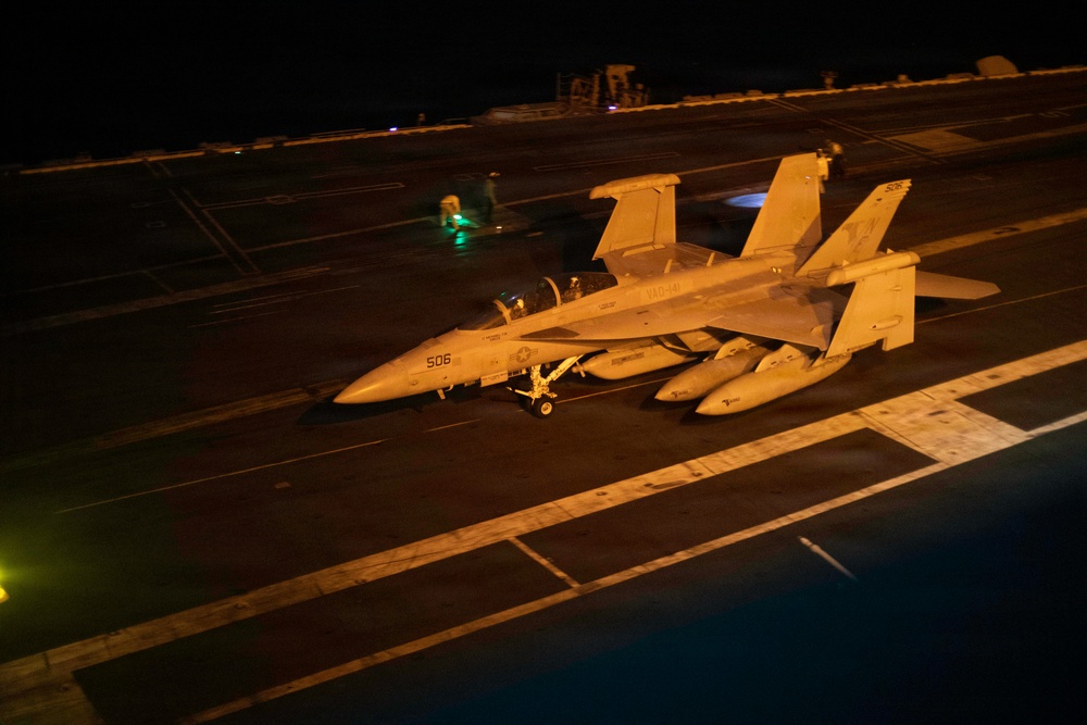 A U.S. Navy EA-18G Growler electronic warfare aircraft, marked with bureau number 506 and squadron designation VAQ-141, is positioned on an aircraft carrier flight deck at night. The aircraft is loaded with external stores including what appear to be fuel tanks and electronic jamming pods. The scene is illuminated by artificial deck lighting, with flight deck personnel visible in the background.
