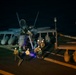 A nighttime scene on what appears to be a naval aircraft carrier flight deck, showing military personnel gathered around a twin-engine jet aircraft, likely an F/A-18 Hornet or Super Hornet. Ground crew members in various colored jerseys are conducting pre-flight or post-flight operations around the aircraft. At least one person appears to be climbing into or out of the cockpit via a ladder.