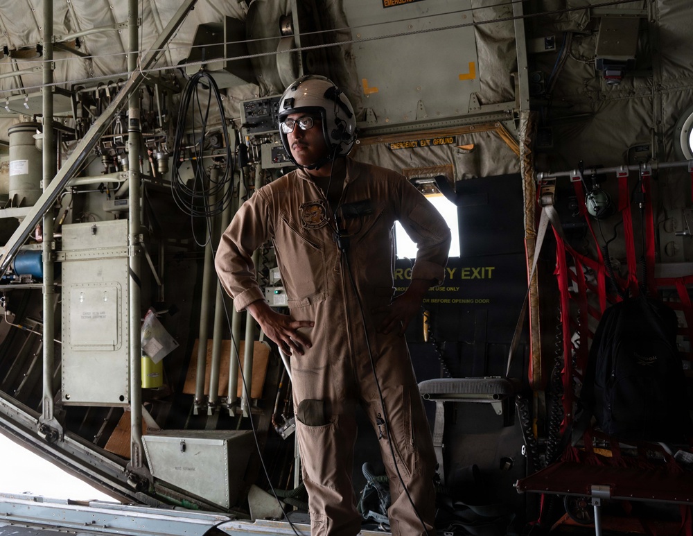 A military aircrew member in tan/khaki flight suit stands inside the cargo bay of a large military transport aircraft, likely a C-130 Hercules. The individual is wearing a flight helmet with communications equipment and protective eyewear, standing near the rear ramp area. The interior shows complex mechanical systems, wiring, and safety markings typical of military cargo aircraft.