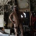 A military aircrew member in tan/khaki flight suit stands inside the cargo bay of a large military transport aircraft, likely a C-130 Hercules. The individual is wearing a flight helmet with communications equipment and protective eyewear, standing near the rear ramp area. The interior shows complex mechanical systems, wiring, and safety markings typical of military cargo aircraft.