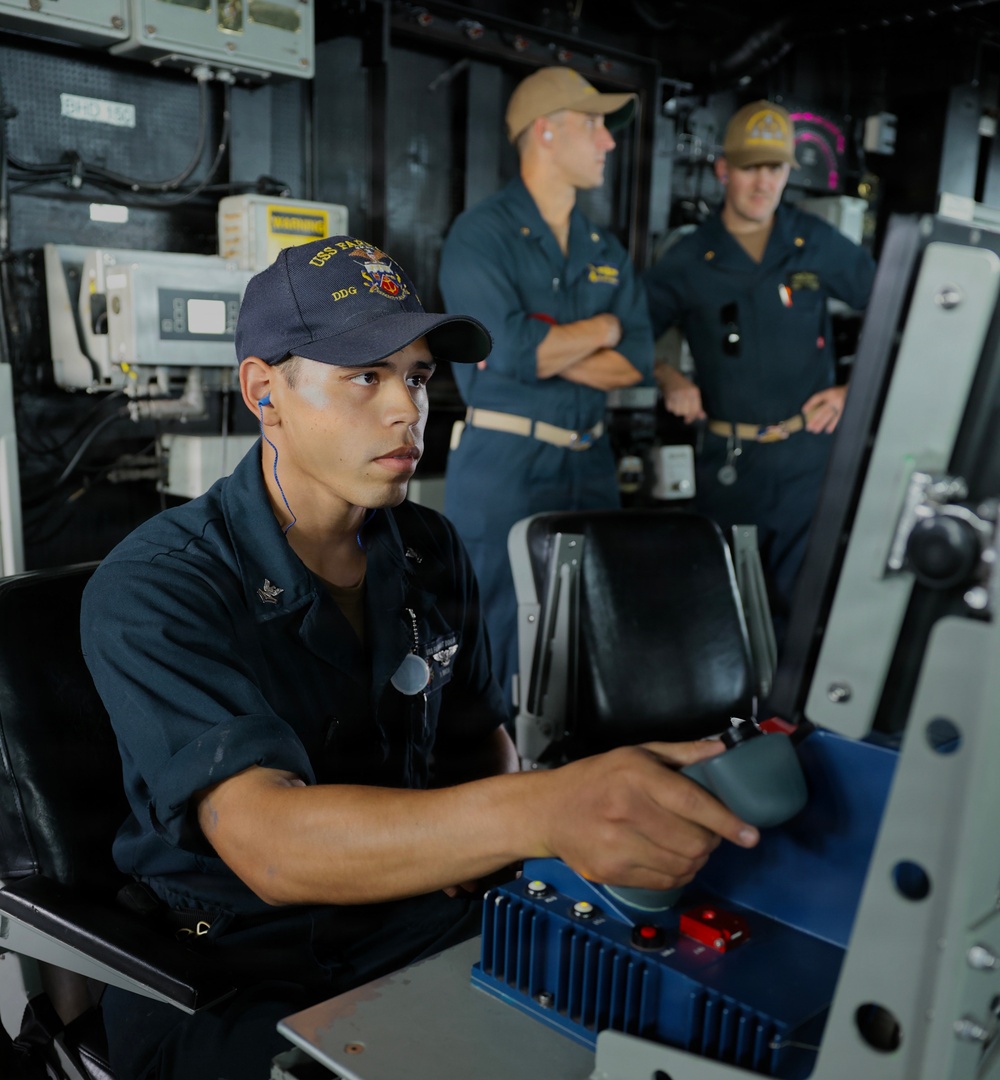 Three U.S. Navy personnel are shown inside what appears to be the bridge or combat information center of a naval vessel. The foreground sailor, wearing a cap marked 'USS' and 'DDG', is seated at a control station operating a joystick or helm control. Two other sailors in the background, wearing tan/khaki caps, appear to be observing or supervising.
