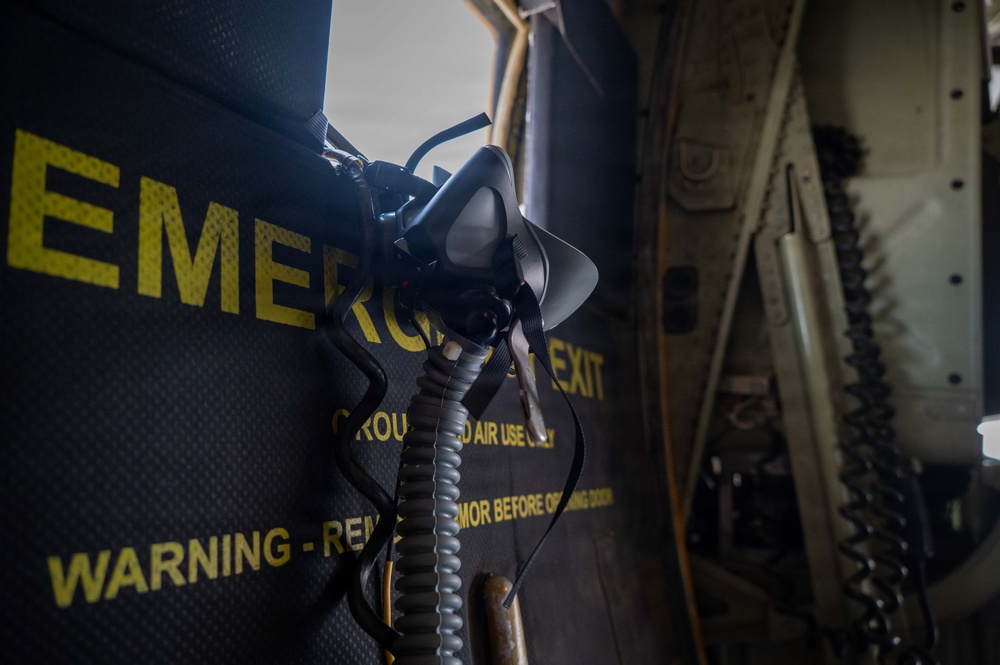 The image shows the interior of a military or large transport aircraft, focusing on an emergency exit door with yellow warning text and a hanging oxygen/breathing mask with a coiled hose. The dark panel displays partial text including 'EMERGENCY EXIT', 'GROUND AIR USE ONLY', and 'WARNING - REMOVE ARMOR BEFORE OPENING DOOR'. The composition is a close-up, low-light shot emphasizing safety equipment mounted near the exit.