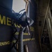 The image shows the interior of a military or large transport aircraft, focusing on an emergency exit door with yellow warning text and a hanging oxygen/breathing mask with a coiled hose. The dark panel displays partial text including 'EMERGENCY EXIT', 'GROUND AIR USE ONLY', and 'WARNING - REMOVE ARMOR BEFORE OPENING DOOR'. The composition is a close-up, low-light shot emphasizing safety equipment mounted near the exit.