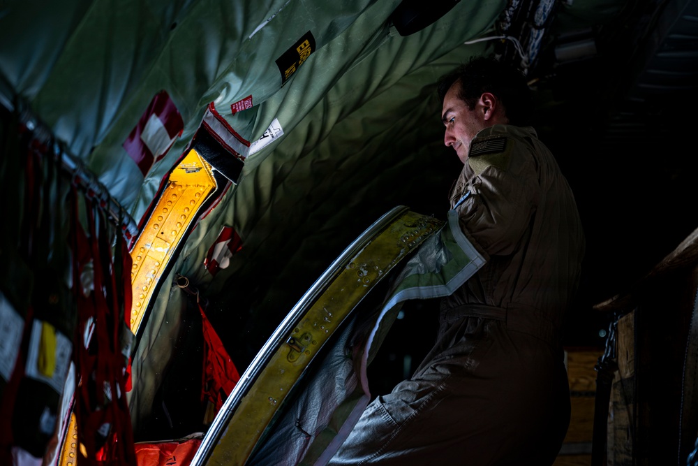 A military aircrew member in an olive drab flight suit is inspecting or operating equipment inside what appears to be a military transport aircraft. The individual is looking toward an open hatch or door area illuminated by yellow interior lighting. The scene is dimly lit with dramatic contrast between shadow and light, suggesting a nighttime or low-light operational environment.