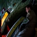 A military aircrew member in an olive drab flight suit is inspecting or operating equipment inside what appears to be a military transport aircraft. The individual is looking toward an open hatch or door area illuminated by yellow interior lighting. The scene is dimly lit with dramatic contrast between shadow and light, suggesting a nighttime or low-light operational environment.