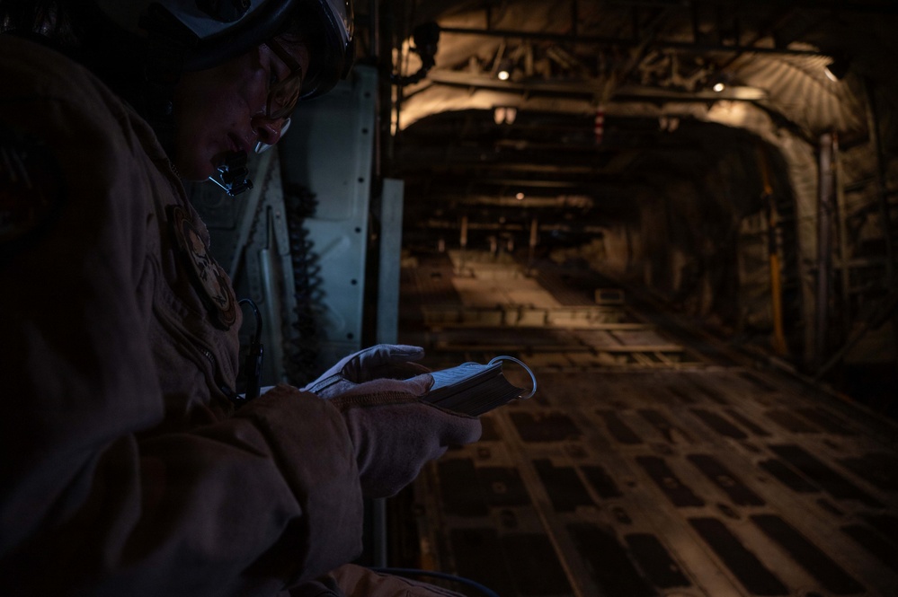 A military aircrew member in flight gear and helmet is reading a small notebook or document inside the cargo hold of a large military transport aircraft. The interior is dimly lit, revealing the characteristic ribbed fuselage structure and cargo floor tie-down rails of what appears to be a C-17 Globemaster or similar heavy-lift transport. The scene suggests an in-flight operational or pre-mission review.