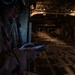 A military aircrew member in flight gear and helmet is reading a small notebook or document inside the cargo hold of a large military transport aircraft. The interior is dimly lit, revealing the characteristic ribbed fuselage structure and cargo floor tie-down rails of what appears to be a C-17 Globemaster or similar heavy-lift transport. The scene suggests an in-flight operational or pre-mission review.
