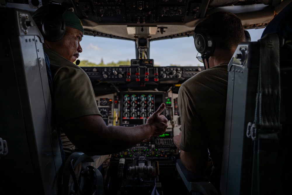 Two crew members are seated in the cockpit of a large military or transport aircraft, engaged in what appears to be a briefing or pre-flight discussion. The individual on the left, wearing a green cap and headset with a boom microphone, is gesturing with one finger toward the instrument panel or the other crew member. The cockpit features an extensive array of analog gauges, switches, and illuminated indicators consistent with a large military transport aircraft.