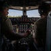 Two crew members are seated in the cockpit of a large military or transport aircraft, engaged in what appears to be a briefing or pre-flight discussion. The individual on the left, wearing a green cap and headset with a boom microphone, is gesturing with one finger toward the instrument panel or the other crew member. The cockpit features an extensive array of analog gauges, switches, and illuminated indicators consistent with a large military transport aircraft.