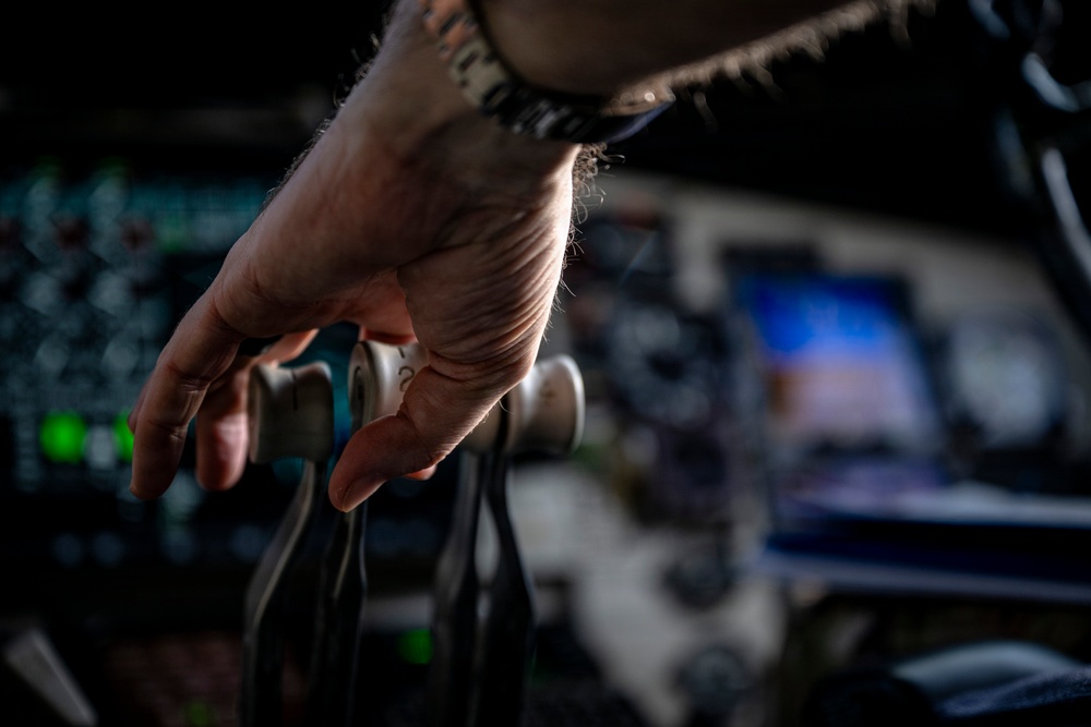 A close-up photograph of a human hand gripping or resting on what appears to be mechanical or rigging equipment with pulleys and cables. The image is dramatically lit with a shallow depth of field, keeping the hand in sharp focus while the background remains blurred. The background suggests a large indoor venue or technical environment with screens and equipment visible.