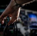 A close-up photograph of a human hand gripping or resting on what appears to be mechanical or rigging equipment with pulleys and cables. The image is dramatically lit with a shallow depth of field, keeping the hand in sharp focus while the background remains blurred. The background suggests a large indoor venue or technical environment with screens and equipment visible.