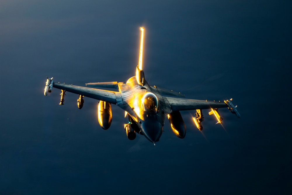 A military fighter jet, consistent with an F-16 Fighting Falcon, is photographed in flight against a dark blue sky. The aircraft is dramatically illuminated by low-angle sunlight, likely at sunrise or sunset, casting intense golden reflections off the canopy, fuselage, and external stores. The jet appears fully armed with multiple wing-mounted munitions and fuel tanks.