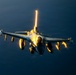 A military fighter jet, consistent with an F-16 Fighting Falcon, is photographed in flight against a dark blue sky. The aircraft is dramatically illuminated by low-angle sunlight, likely at sunrise or sunset, casting intense golden reflections off the canopy, fuselage, and external stores. The jet appears fully armed with multiple wing-mounted munitions and fuel tanks.