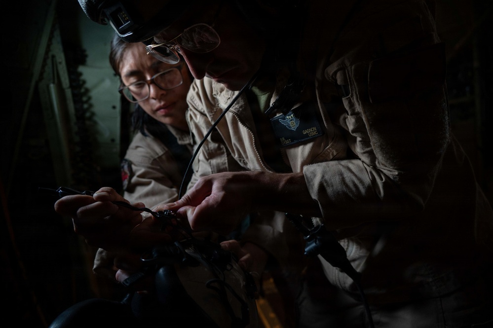 Two military personnel in tan/khaki flight suits are examining a small object, likely a micro drone or small device, in what appears to be the interior of a military aircraft. The foreground individual has a name tape reading 'GAGNON' and 'USMC' with a visible wing insignia patch, indicating United States Marine Corps affiliation. The image is dramatically lit with strong shadows, emphasizing the focused activity.