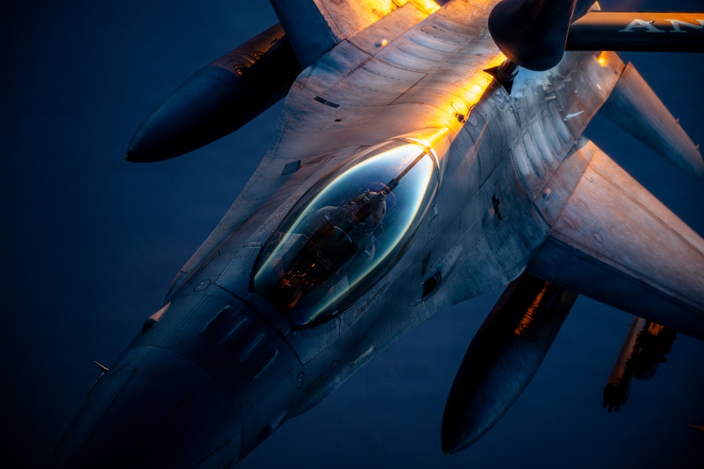 An aerial photograph taken from a tanker aircraft showing a fighter jet, consistent with an F-16 Fighting Falcon, conducting an aerial refueling operation. The image is captured at dusk or dawn, with warm orange lighting illuminating the underside of the tanker's wing and the refueling boom. A pilot is visible in the cockpit of the receiver aircraft.
