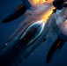 An aerial photograph taken from a tanker aircraft showing a fighter jet, consistent with an F-16 Fighting Falcon, conducting an aerial refueling operation. The image is captured at dusk or dawn, with warm orange lighting illuminating the underside of the tanker's wing and the refueling boom. A pilot is visible in the cockpit of the receiver aircraft.