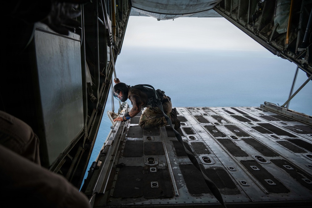 A military personnel member in tactical gear is crouched at the edge of an open cargo ramp of a military transport aircraft, likely a C-130 Hercules, preparing for a jump over a large body of water visible below. The image is taken from inside the aircraft looking outward, capturing the dramatic perspective of the open ramp against the ocean backdrop. The scene suggests a military airborne or special operations training exercise.