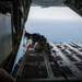 A military personnel member in tactical gear is crouched at the edge of an open cargo ramp of a military transport aircraft, likely a C-130 Hercules, preparing for a jump over a large body of water visible below. The image is taken from inside the aircraft looking outward, capturing the dramatic perspective of the open ramp against the ocean backdrop. The scene suggests a military airborne or special operations training exercise.