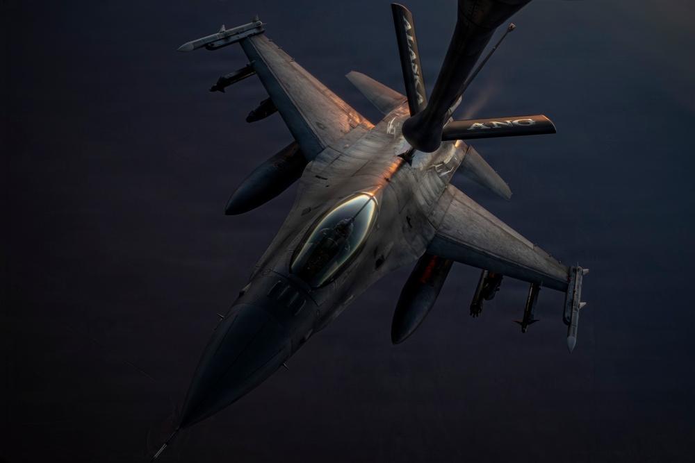An F-16 Fighting Falcon jet fighter is captured in a dramatic aerial refueling operation, receiving fuel from a tanker aircraft via a boom visible at the top of the frame. The aircraft is photographed from above and slightly forward, revealing its full weapons loadout and cockpit. The image is taken during low-light conditions, with warm golden light illuminating the top surfaces of the aircraft against a dark sky.