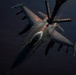 An F-16 Fighting Falcon jet fighter is captured in a dramatic aerial refueling operation, receiving fuel from a tanker aircraft via a boom visible at the top of the frame. The aircraft is photographed from above and slightly forward, revealing its full weapons loadout and cockpit. The image is taken during low-light conditions, with warm golden light illuminating the top surfaces of the aircraft against a dark sky.